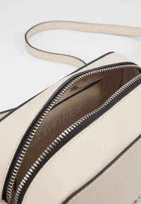 Beżowa skórzana torba na ramię z czarnymi akcentami i srebrnym zamkiem błyskawicznym, z widoczną teksturą i widocznym logo marki w środku.