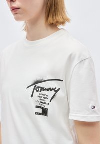Άτομο που φοράει λευκό T-shirt Tommy Jeans με μαύρες εκτυπώσεις ονομάτων πόλεων και λογότυπου στο στήθος και μικρό λογότυπο στο μανίκι.