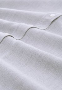 Tessuto grigio chiaro che mostra una texture liscia con morbide onde e un dettaglio di bottone bianco. Il materiale appare leggero e traspirante.
