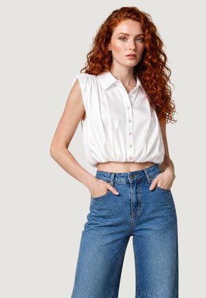 Jeune femme aux longs cheveux roux bouclés porte un crop top blanc sans manches boutonné et un jean bleu taille haute, debout les mains dans les poches.