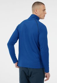 Modré športové tričko s dlhým rukávom a vysokým golierom, textúrovaná tkanina, kontrastné čierne švy a voľný strih, nosené s tmavými nohavicami.