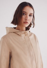 Giacca beige con cappuccio realizzata in tessuto liscio, dotata di due chiusure a bottone e colletto alto, con cuciture minime per un design pulito.