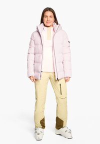 Giacca puffa rosa chiaro con colletto alto, zip beige e strato sottile. Pantaloni da sci beige con tasche con zip e accenti marroni.