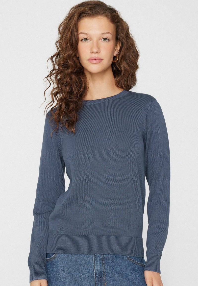 Donna con capelli ricci castani indossa un maglione blu a maniche lunghe semplice e jeans blu, in piedi davanti a uno sfondo chiaro e uniforme.