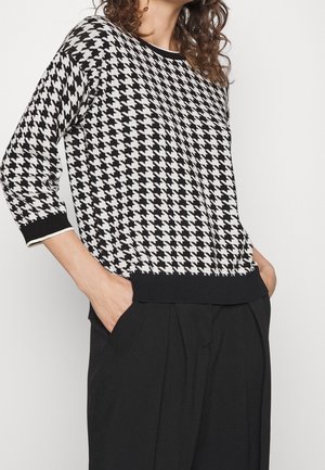 Femme portant un pull à motif pied-de-poule noir et blanc à manches trois-quarts et un pantalon noir, les mains dans les poches.