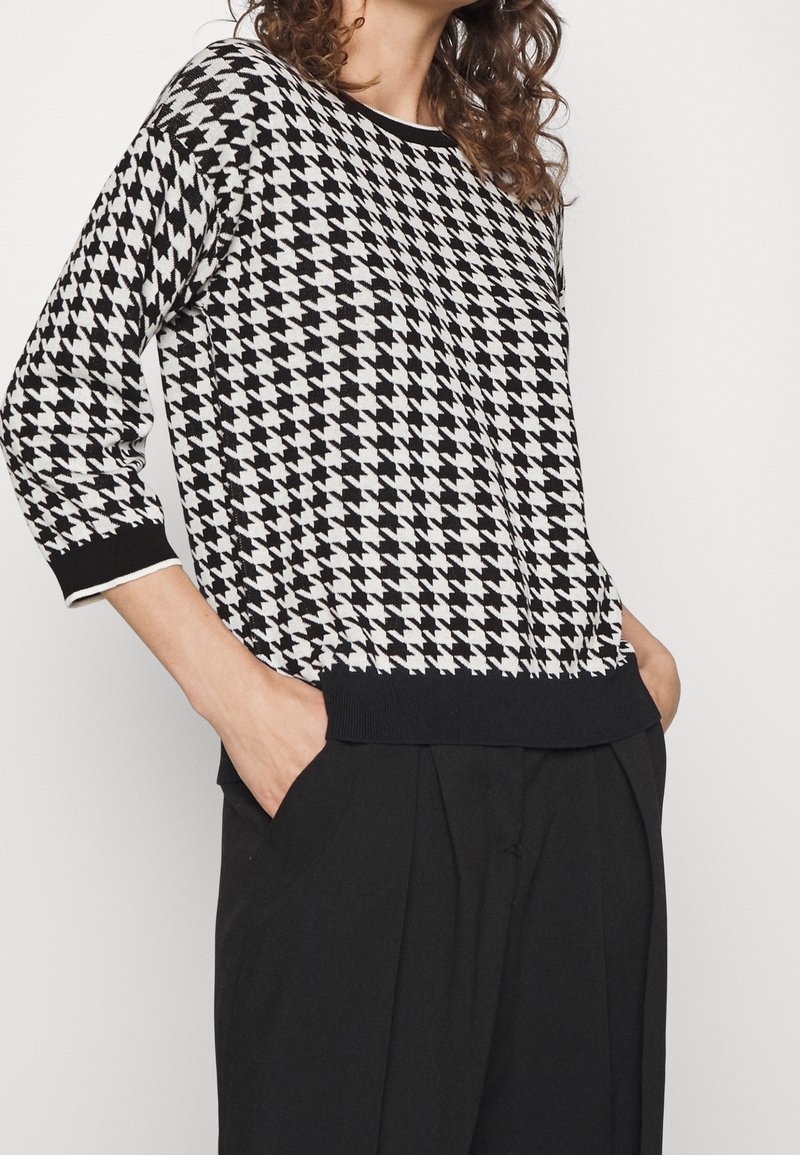 Femme portant un pull à motif pied-de-poule noir et blanc à manches trois-quarts et un pantalon noir, les mains dans les poches.