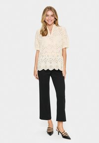 Cremfarbene Bluse mit Augenmuster, kurzen Ärmeln, gewelltem Saum und V-Ausschnitt, kombiniert mit schwarzen Hosen und schwarzen Riemchen-Heels.