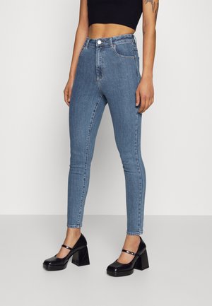 Person wearing high-waisted light blue skinny jeans, black cropped top, and black Mary Jane platform heels, standing against plain light background.