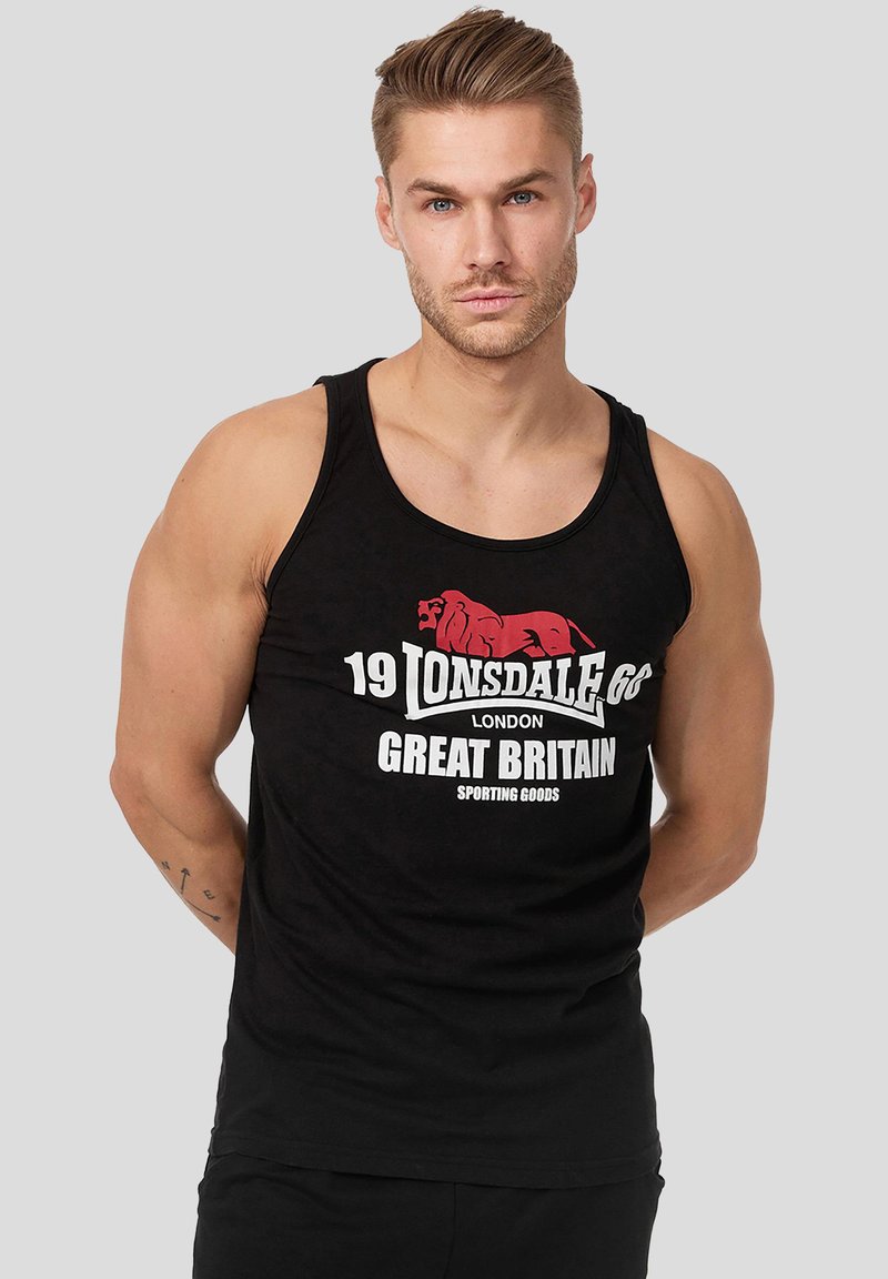 Homme athlétique aux cheveux coiffés portant un débardeur noir Lonsdale London Great Britain, debout les mains derrière le dos sur fond gris.