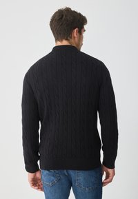 Czarny sweter w splot łańcuszkowym z ribbowanym kołnierzykiem i mankietami, z teksturowanym wzorem. Noszony z niebieskimi dżinsami, prezentując luzny krój.