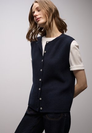 Gilet tricoté bleu marine à col rond, fermeture à boutons et bordures côtelées, porté par-dessus un t-shirt blanc, complété par un jean foncé.