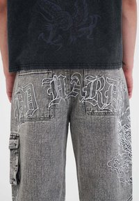 Szare dżinsowe spodnie z kontrastowym białym haftowanym tekstem i wzorami. Posiadają tylne kieszenie oraz kieszeń typu cargo z boku.