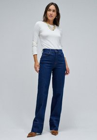 Jean en denim bleu foncé taille haute avec des jambes droites et des coutures visibles, associés à un haut blanc à manches longues et des chaussures pointues marron.