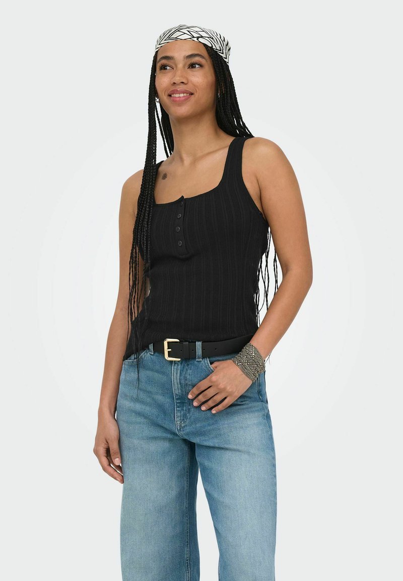 Femme avec de longues tresses portant un débardeur noir, un jean bleu clair avec une ceinture noire, un foulard à motifs et un bracelet en argent, souriant légèrement.