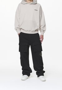Beige huvtröja med framfickor och logotyp, kombinerad med lösa svarta cargobyxor med flera fickor och ljusa accenter. Vita sneakers.