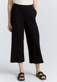 Pantalons larges noirs fabriqués dans un tissu léger et doux ; dotés d'une taille élastique et de poches latérales. Assortis avec des sandales noires.