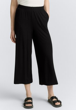 Pantalons larges noirs fabriqués dans un tissu léger et doux ; dotés d'une taille élastique et de poches latérales. Assortis avec des sandales noires.