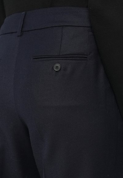 Tamno plave hlače glatke teksture, s jednim stražnjim džepom i detaljem jednog crnog gumba. Krojački dizajn bez vidljivih ukrasa.
