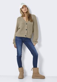 Beige neulecardigan, jossa on V-pääntie ja mustat napit, yhdistettynä sinisiin tiukkoihin farkkuihin ja pehmeän, pörröisen tekstuurin omaaviin vaaleanruskeisiin nilkkureihin.