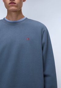 Blå genser med rund hals laget av mykt materiale, med et lite brodert norsk flagg på brystet og kontrasterende lysfarget halslinning.