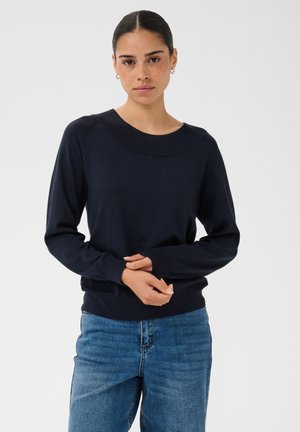 Femme aux cheveux foncés attachés, portant des boucles d’oreilles créoles argentées, un pull bleu marine à manches longues et un jean bleu sur un fond clair uni.