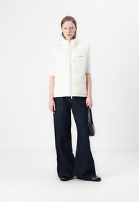 Hvit polstret vest med høy krage, glidelås foran og logodetalj. Kombinert med mørke, vide jeans og svarte, flate sandaler med åpen tå.