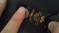 Zwarte stoffen label met oranje tekst, zilveren metalen knop met de tekst "ALPHA," zorgsymbolen en logo. Vinger raakt het label aan.