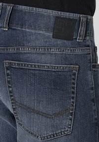 Dunkelblaue Denim-Jeans verfügen über eine strukturierte Oberfläche, einen geraden Schnitt und hintere Taschen mit markanten Nähten. Das Etikett am Bund ist schwarz.