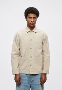 Beżowa kurtka z płótna z zapięciem na guziki z przodu, ostrym kołnierzem i dwoma kieszeniami na piersi. Charakteryzuje się luźnym krojem i widocznymi detalami przeszyć.