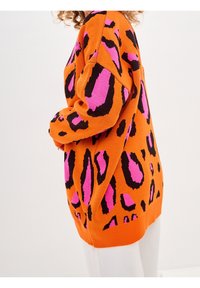 Oranžový pletený svetr s černo-růžovým leopardím vzorem, s volnějším střihem a žebrovanými manžetami. Materiál je měkký a strukturovaný.