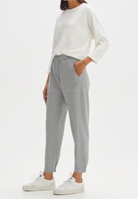 Femme portant un haut blanc à manches longues et un pantalon gris clair à taille élastique avec des poches latérales, ainsi que des baskets blanches, debout avec une main dans une poche.