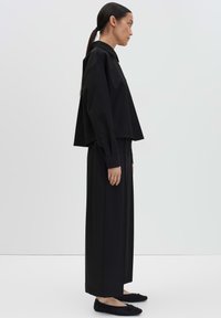 Vrouw in profiel staande, gekleed in een losse zwarte blouse met lange mouwen, wijd zwarte broek en zwarte platte schoenen voor een effen witte achtergrond.