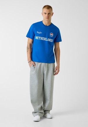 Jeune homme portant un T-shirt bleu "Pays-Bas", pantalon de survêtement large gris et baskets blanches, debout devant un fond blanc uni.