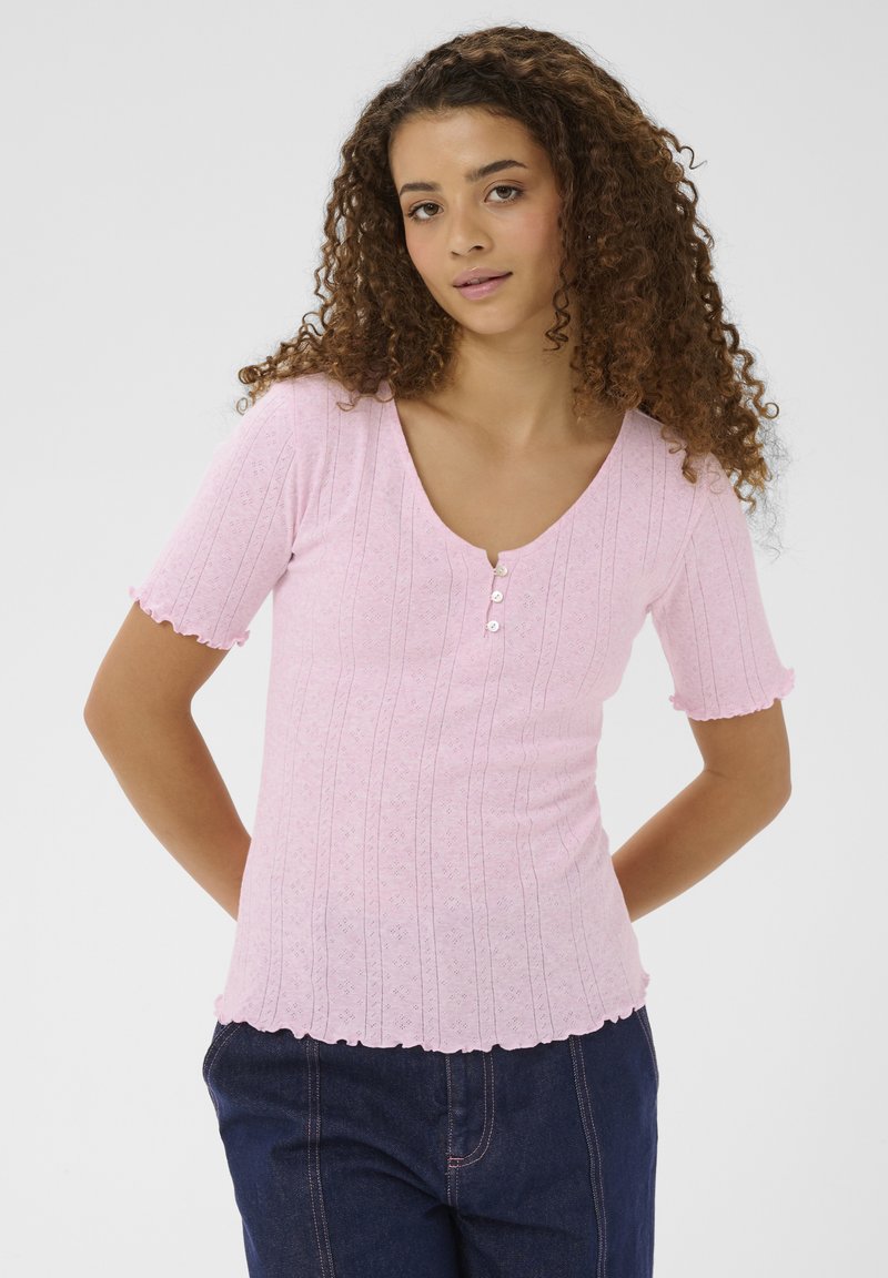 Mlada žena kovrčave kose u svijetloružičastoj ribanoj majici s kratkim rukavima i gumbima, u kombinaciji s tamnoplavim trapericama.