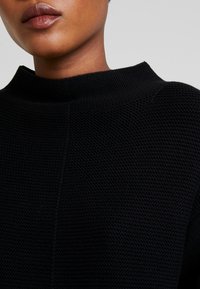 Haut en tricot noir texturé avec un col montant et des détails de couture minimaux, mettant en avant un design simple et moderne.