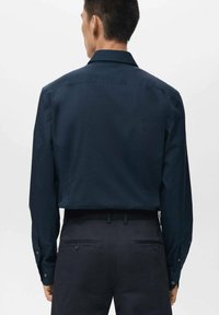 Camisa de manga larga azul oscuro con un cuello estructurado y detalles de botones, combinada con pantalones oscuros. Se aprecia la textura suave de la tela.