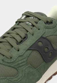 Maslinasto zelena suede i tekstilna tenisica s tamnozelenim naglascima, teksturiranim jezikom, okruglim čipkama i crnim logotipom na bočnoj strani.