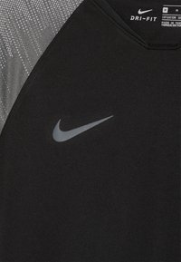 Camiseta deportiva negra Nike Dri-FIT con panel de hombro gris con patrón. Presenta una textura suave y un logotipo Nike gris reflectante.