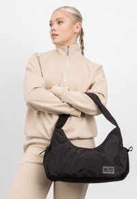 Schwarze Nylon-Schultertasche mit einer geschwungenen Form, Reißverschlussverschluss und einem Logopatch aus Stoff. Der Gurt ist breit und gepolstert für zusätzlichen Komfort.