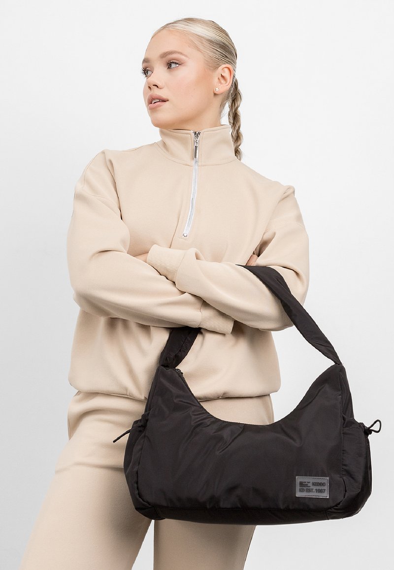 Schwarze Nylon-Schultertasche mit einer geschwungenen Form, Reißverschlussverschluss und einem Logopatch aus Stoff. Der Gurt ist breit und gepolstert für zusätzlichen Komfort.