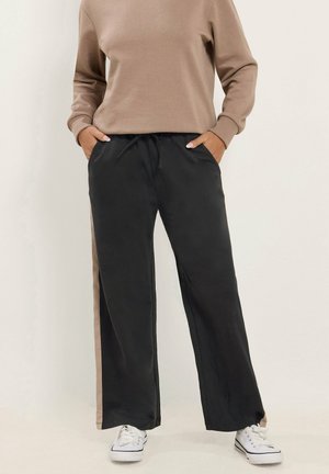 Person wearing black wide-leg pants with beige side stripes, beige long-sleeve top, and white sneakers, standing with hands in pockets.