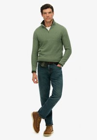 Grüner Strickpullover mit Reißverschlusskragen, kombiniert mit einem karierten Hemd. Trägt man zu blauen Jeans und braunen Schnürstiefeln. Strukturiertes Finish auf dem Stoff.