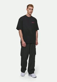 Čierne oversized tričko s krátkymi rukávmi a jemným červeným logom, v kombinácii s tmavými cargo nohavicami a bielymi teniskami. Jednoduchý a funkčný outfit.