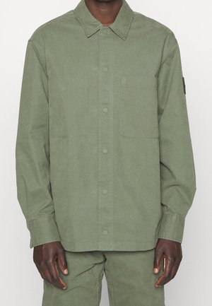 Man wearing olive green button-up shirt with chest pocket and matching pants, standing against plain light background.