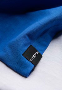 Modrá bavlněná tričko s hladkou texturou a krátkými rukávy, s černou etiketou s nápisem "ombre" na lemu.