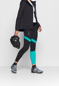 Svart vattentät jacka i kombination med svarta och teal leggings med kontrasterande paneler, tillsammans med grå terrängskor och en svart hjälm.