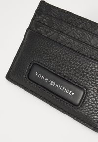 Tarjetero de cuero negro con un patrón texturizado, tres espacios para tarjetas y una placa de logo en relieve con "TOMMY HILFIGER" en blanco.