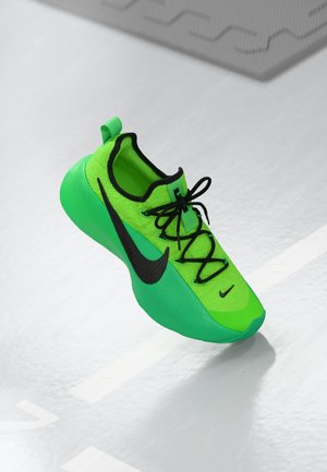 Chaussure de sport vert vif avec des lacets noirs et des logos Nike noirs sur une surface gris clair brillante.
