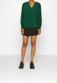 Zielona bluzka z długim rękawem i dekoltem w kształcie litery V, z fałdowanymi ramionami, zestawiona z czarną mini spódnicą i czarnymi mokasynami. Gładka tekstura, casualowy styl.