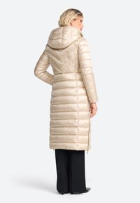 Langer beiger Puffer-Mantel mit Kapuze, der über gestepptes Zickzackmuster und seitliche Reißverschlüsse verfügt. Glatte, glänzende Textur, reicht bis zur Mitte der Wade.